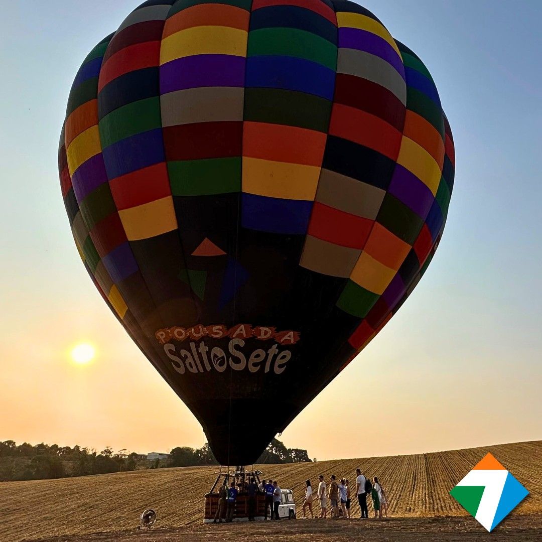 Balão de ar quente com painéis coloridos sobrevoando um campo ao pôr do sol, com pessoas em pé embaixo.