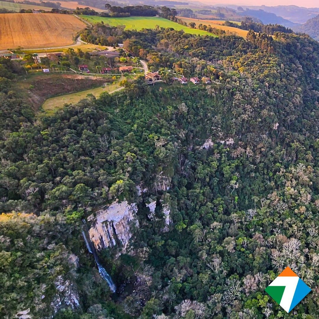 Vista aérea: floresta verdejante, cachoeira na encosta de um penhasco, terras agrícolas ao longe.