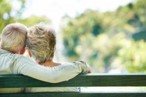 An elderly couple is sitting on a park bench with their arms around each other.