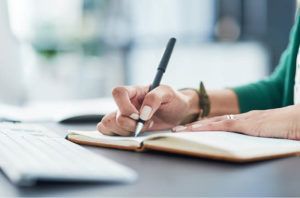 A woman is writing in a notebook while sitting at a desk.