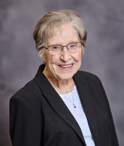 An elderly woman wearing glasses and a black jacket is smiling for the camera.