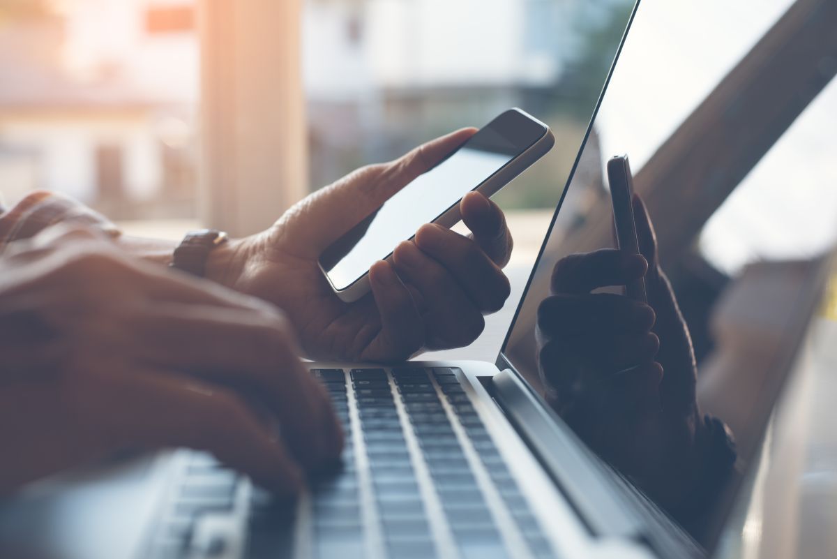 A person is typing on a laptop while holding a cell phone.