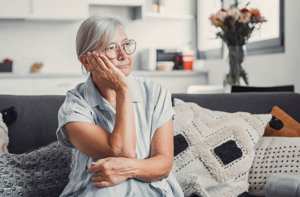 Older woman sitting alone on her couch at home, resting her head on her hand while grieving the loss of a loved one