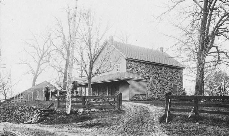 Stone farmhouse with porch, gravel driveway, and wooden fence. Bare trees surround the building.