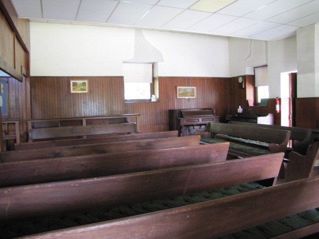 Wooden pews in a church interior with wood paneling, a piano, and two small paintings.