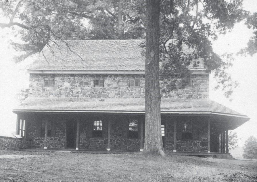 Two-story stone house with wrap-around porch, tall tree in front.