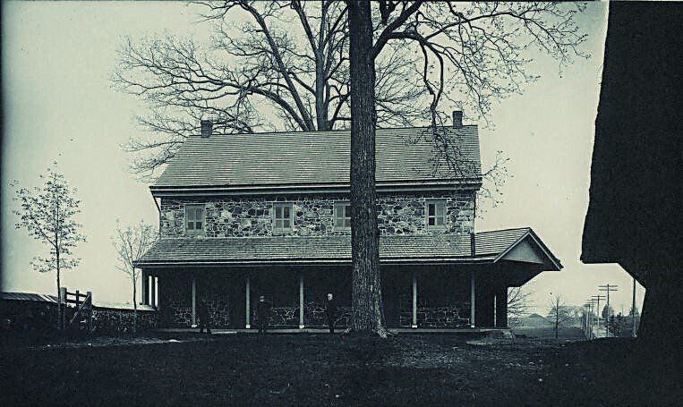 Stone house with porch, leafless tree behind it, in black and white.