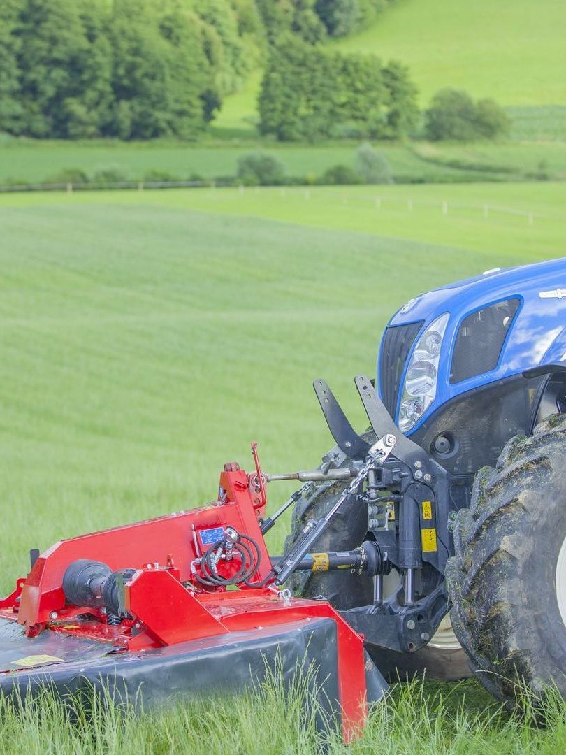 A blue tractor with a red mower attached to it is parked in a grassy field.
