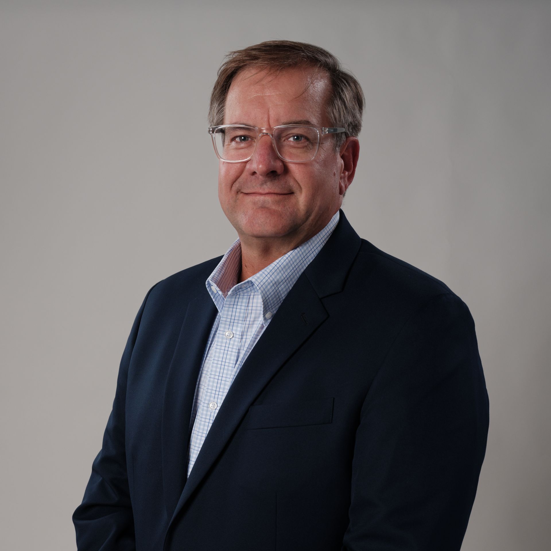 Man in a blue blazer and glasses, smiling slightly, against a gray background.