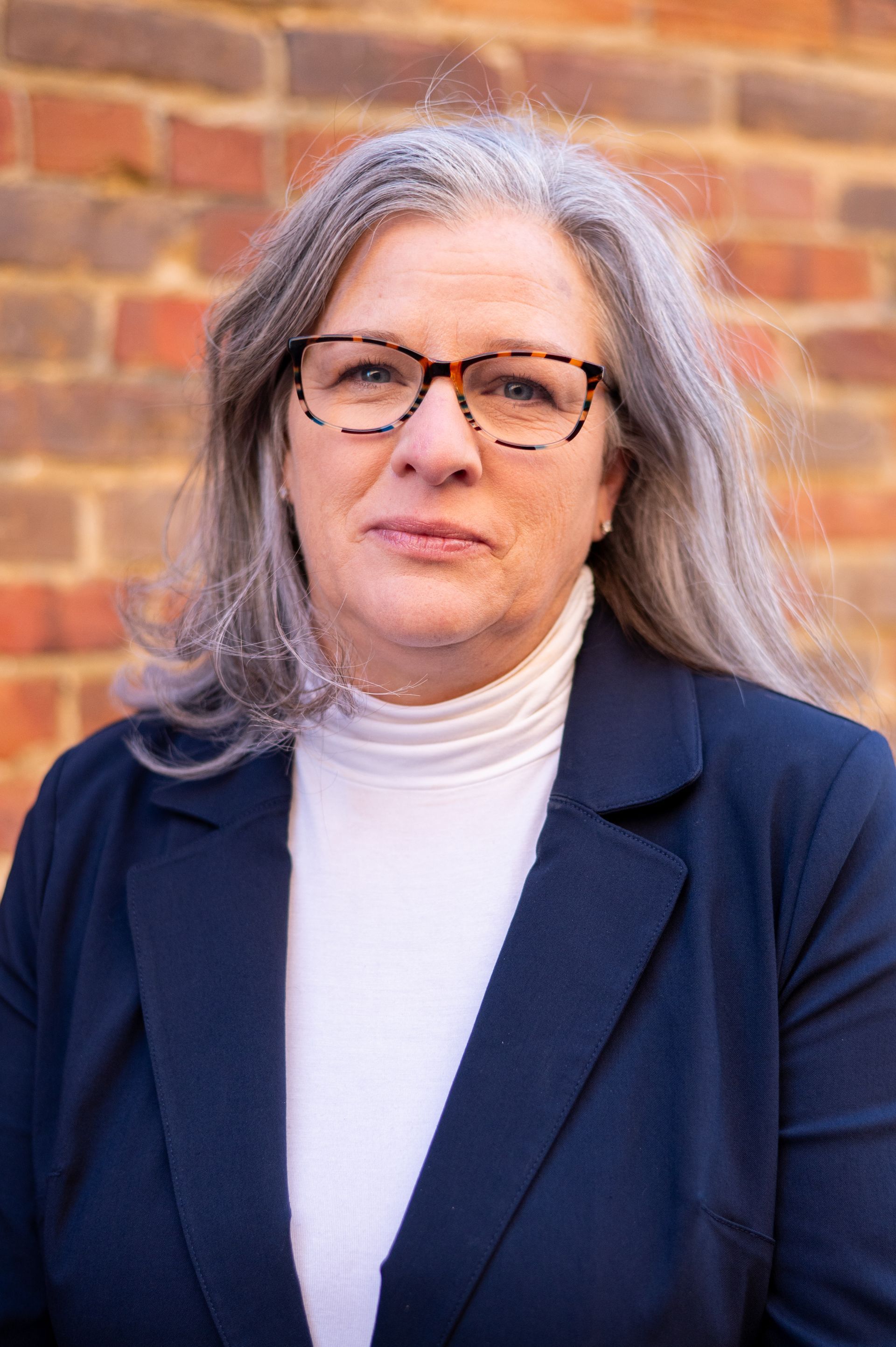Woman with gray hair, glasses, wearing a navy blazer and white turtleneck, in front of a brick wall.