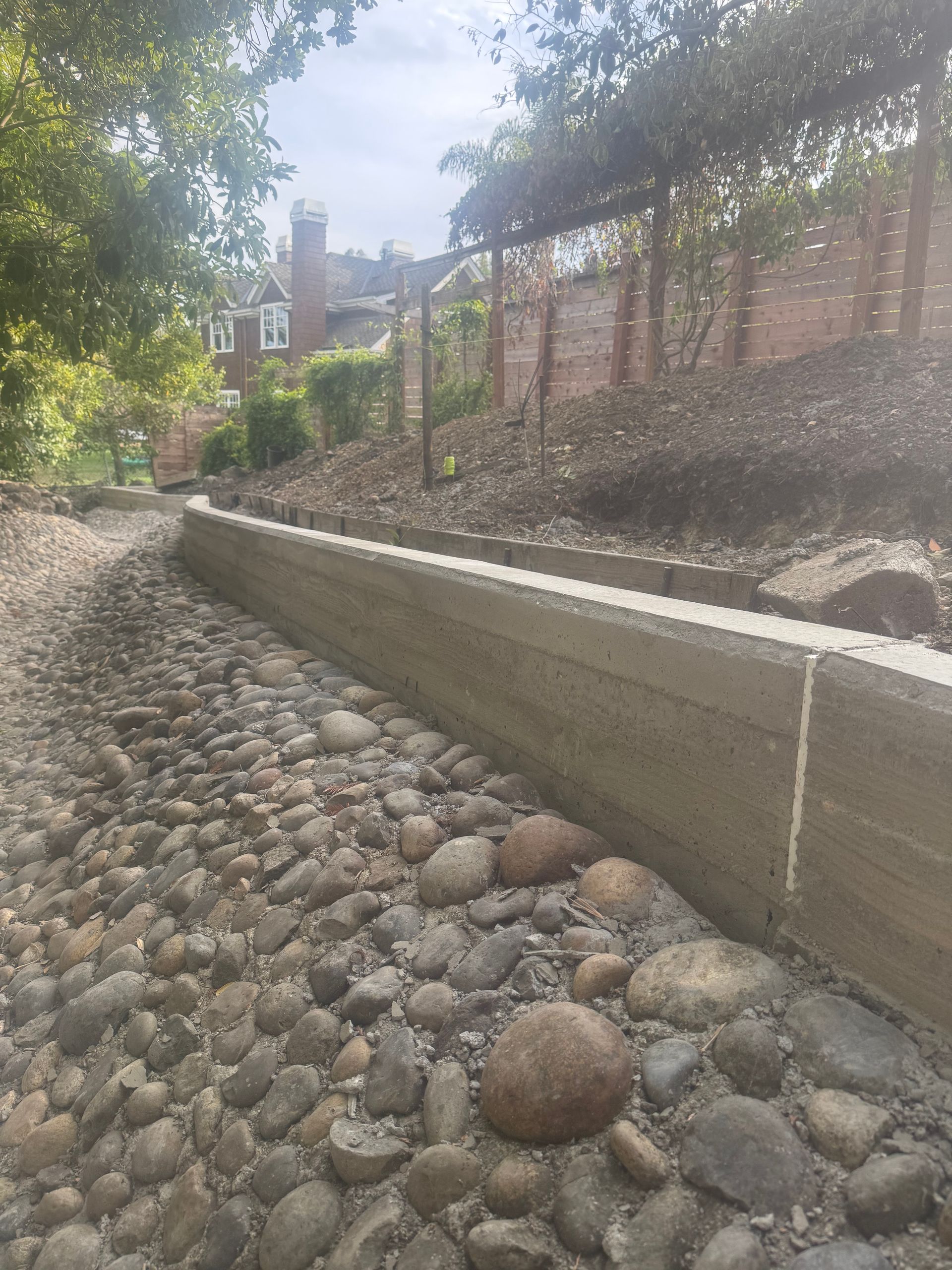 Stone-lined dry creek bed with a concrete retaining wall, leading to a brick building and foliage in the background.