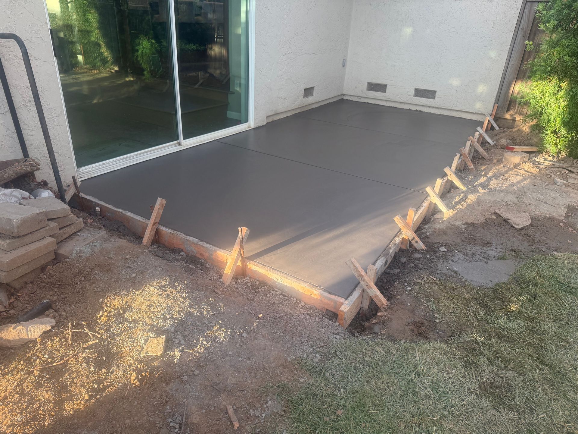 Newly poured concrete patio next to a sliding glass door, framed by wood, surrounded by dirt and grass.