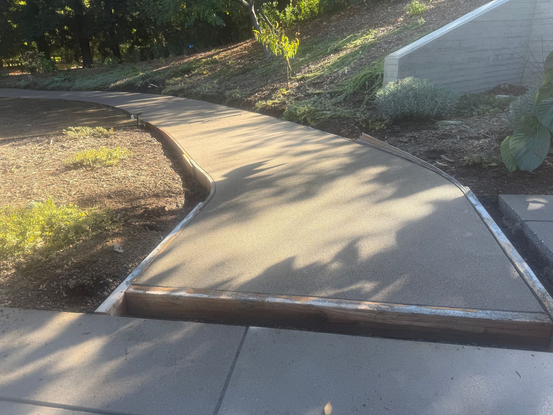 Newly poured concrete walkway curves around a small hill, bordered by dirt and a retaining wall.