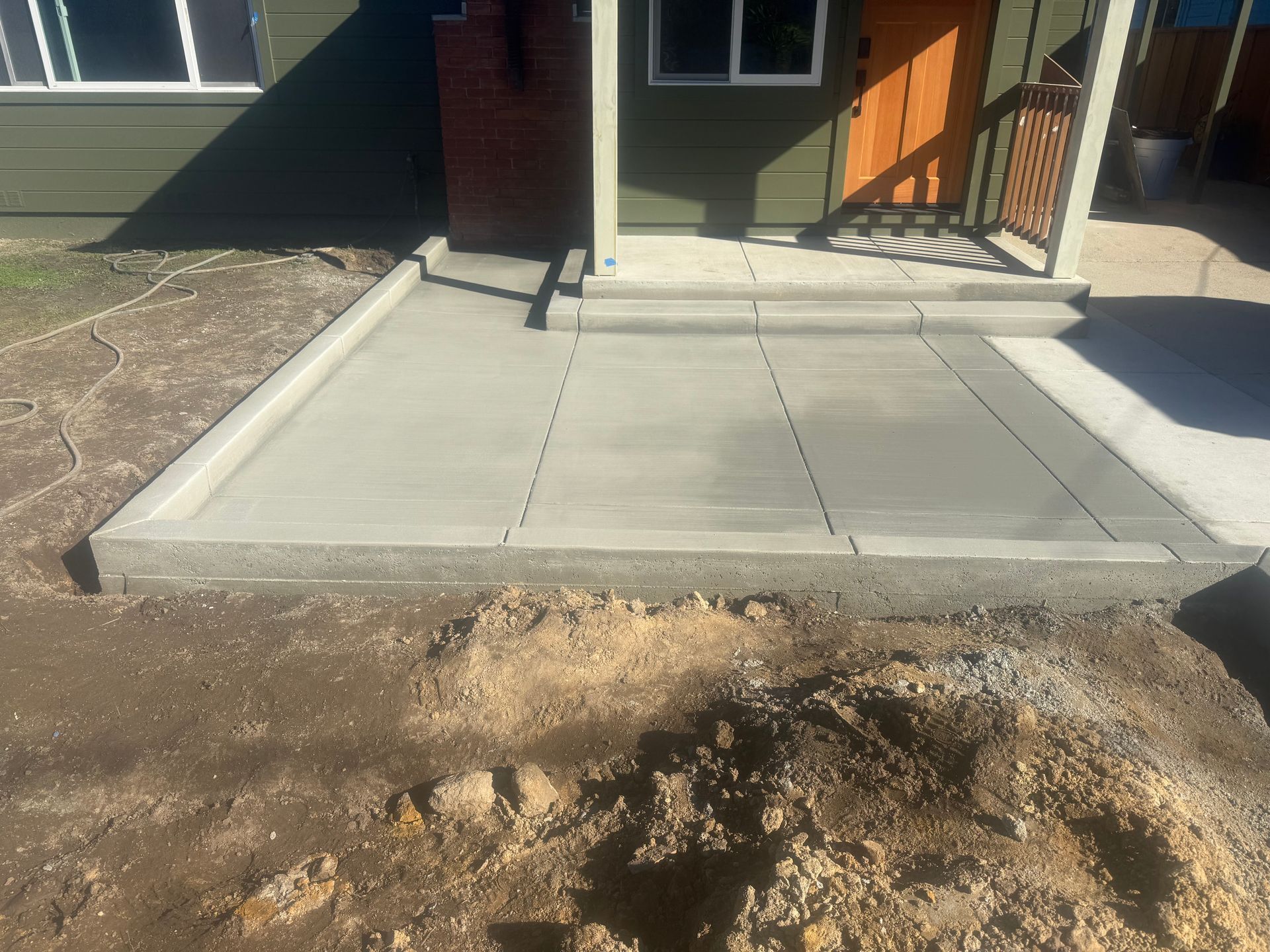 Newly poured concrete porch and steps in front of a green house; dirt in foreground.