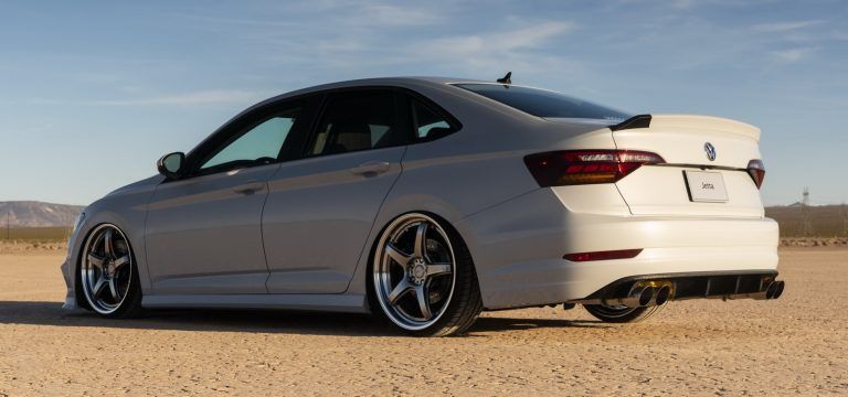 A gray Volkswagen Jetta sedan with custom wheels, lowered stance, and black accents, parked on a dirt road.