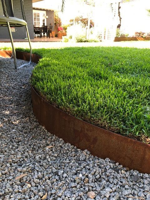 Rusty metal edging borders a lush green lawn, gravel path in foreground. A chair and house in the background.