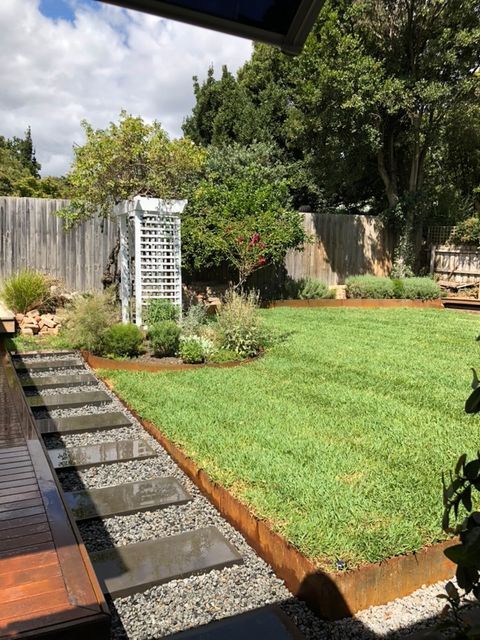 A backyard with a green lawn, stepping stones, wooden deck, and a white trellis.