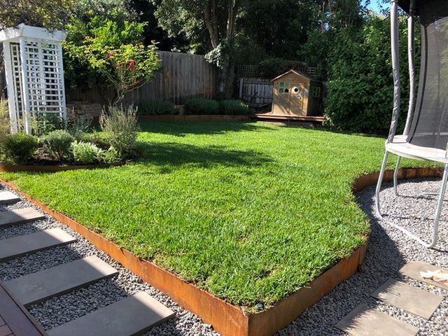 Lush green backyard with a lawn bordered by rusty metal edging, a small wooden playhouse, and a white trellis.