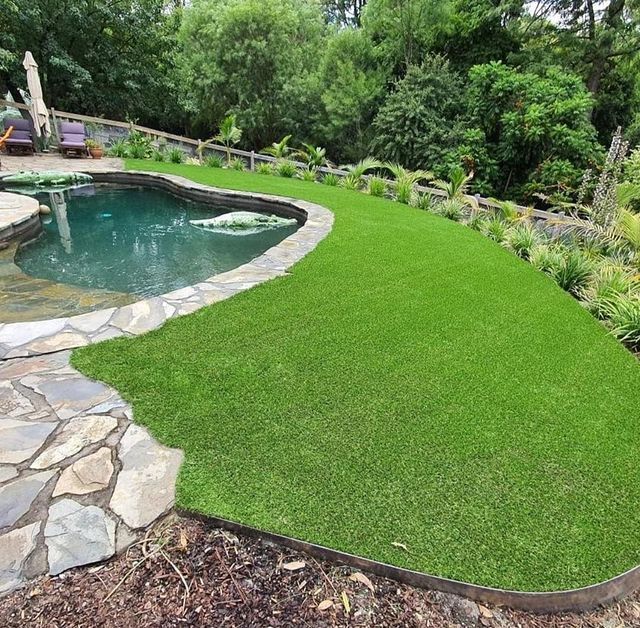 Lush green artificial turf slopes beside a freeform swimming pool with stone coping, in a landscaped backyard.