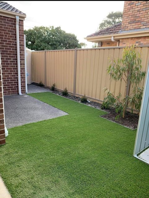 Backyard with artificial green turf, concrete, beige fence, and small plants.