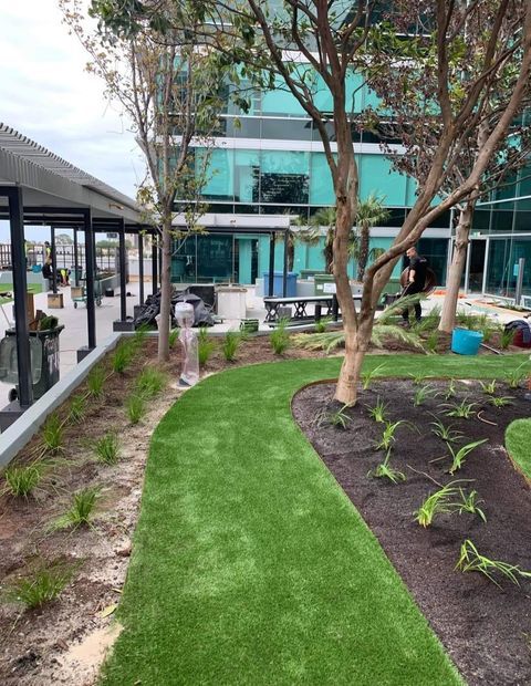 Lush green artificial turf pathway winds through a landscaped courtyard with trees and a building with glass windows.