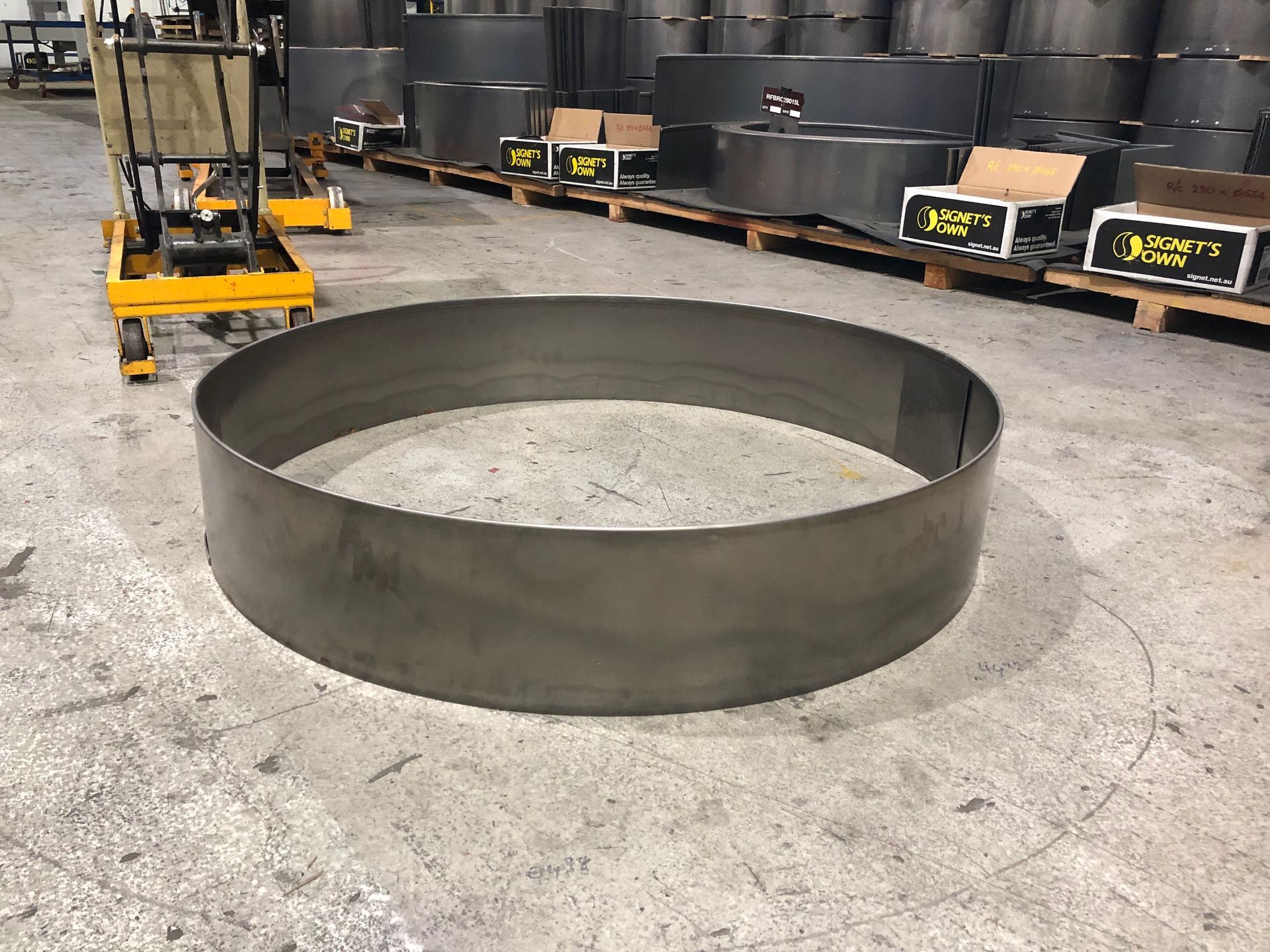 Large, circular metal band on a concrete floor in a warehouse. Other metal cylinders and pallets are visible.