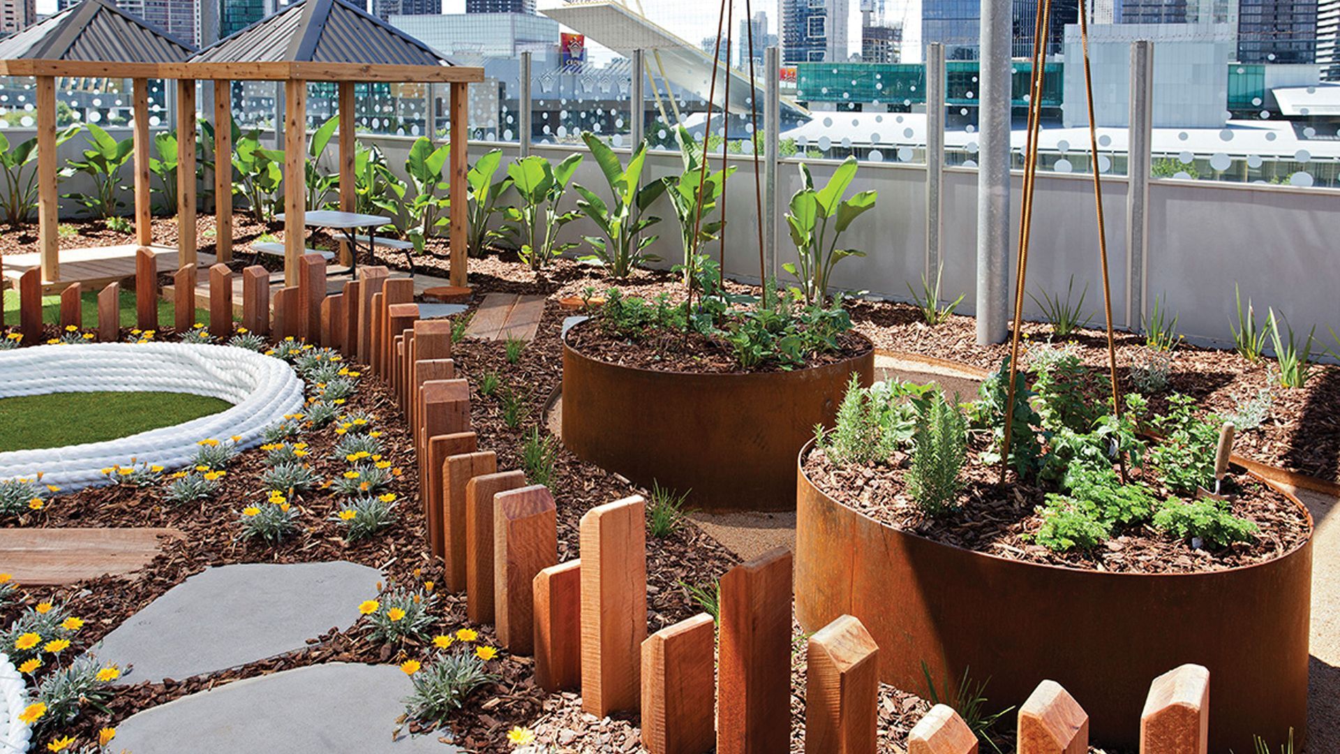 Rooftop garden with raised planters, wooden fence, and pergola. Green plants, gravel, and paving stones are present.