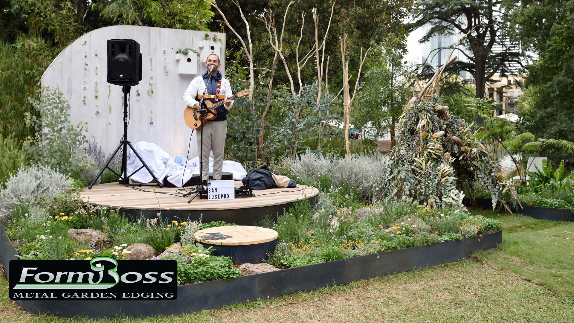 Musician playing guitar on a stage in a garden, with a speaker and backdrop, greenery surrounds.
