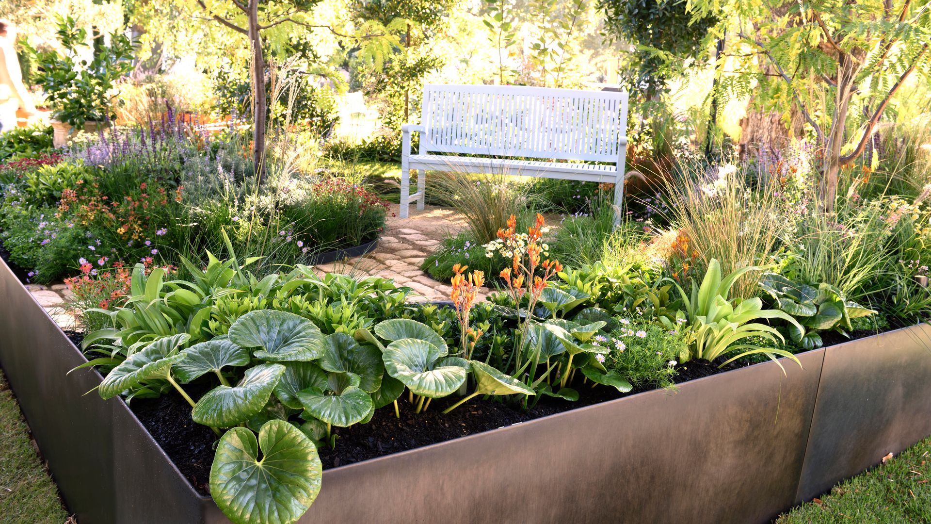 Raised garden bed with lush green plants, a light blue bench, and flowering plants in a sunny garden setting.