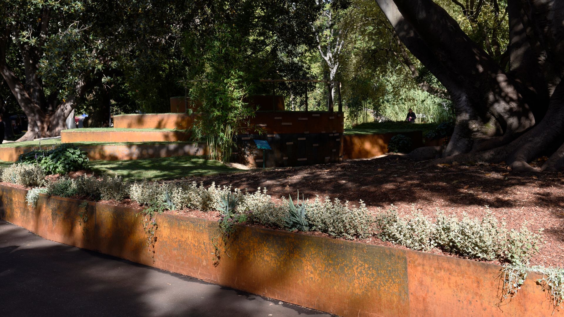 Weathered-steel planters with greenery and steps in a sunny park with large trees.