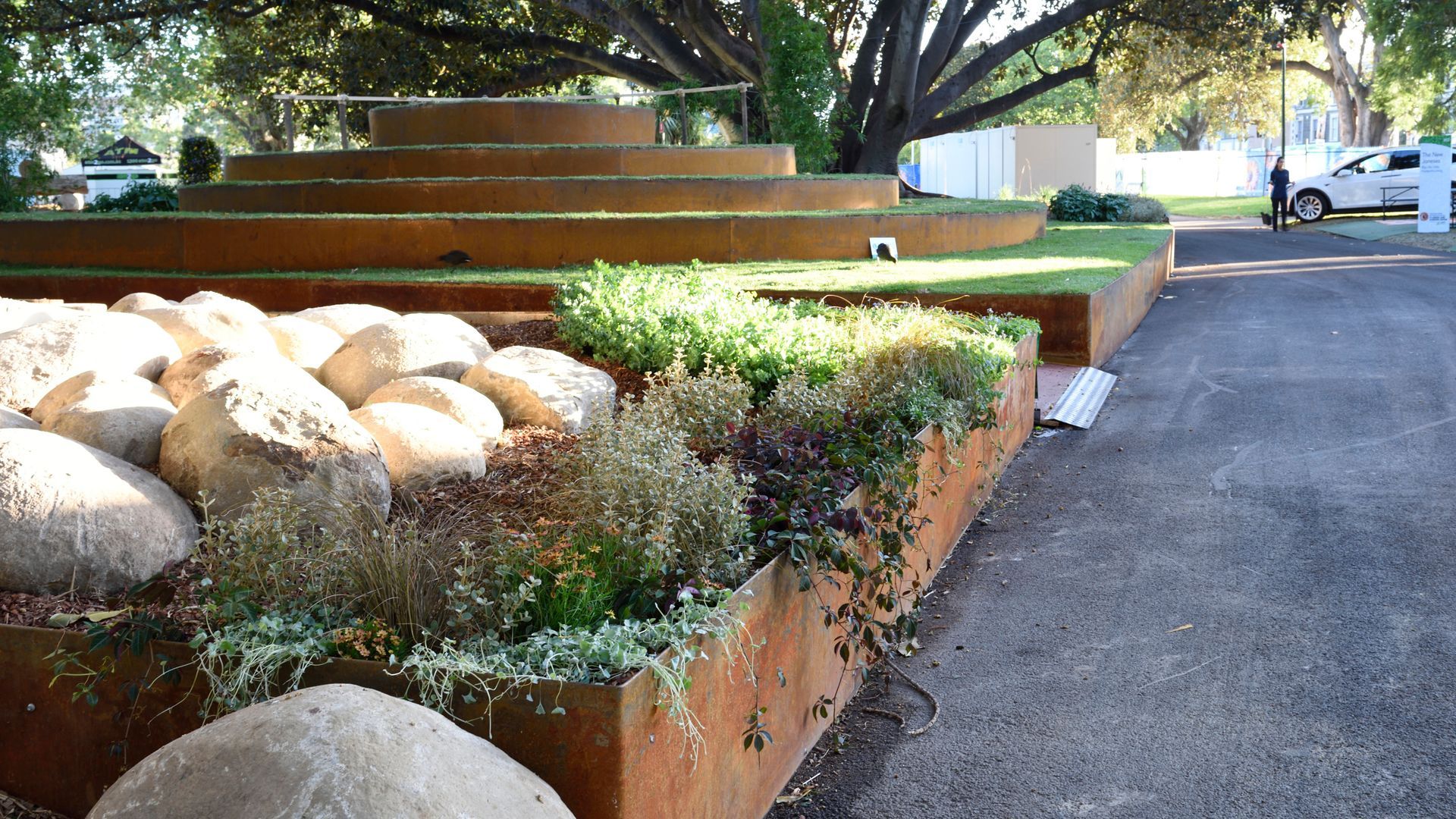 Low-angle view of a tiered, rust-colored steel planter with rocks and various plants next to a paved area.