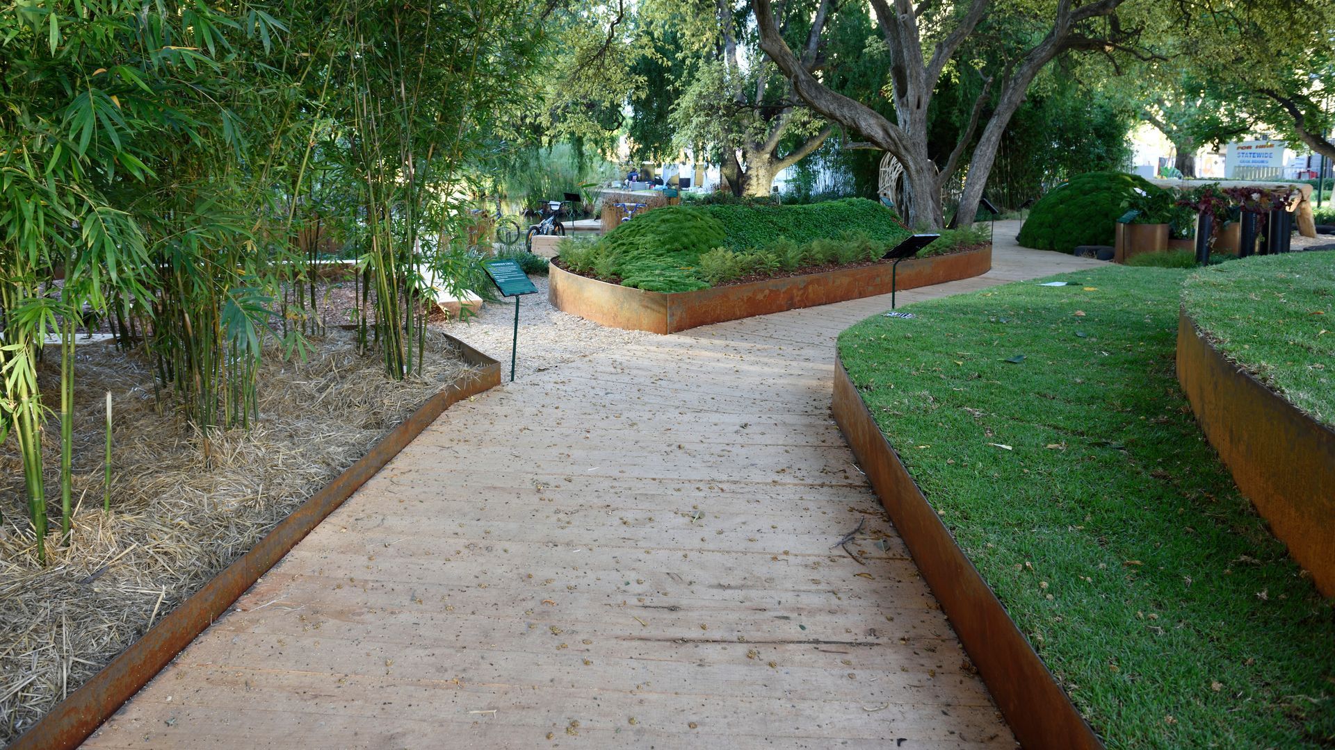 Path with raised grassy beds, framed with rusted metal, in a park-like setting with bamboo and trees.