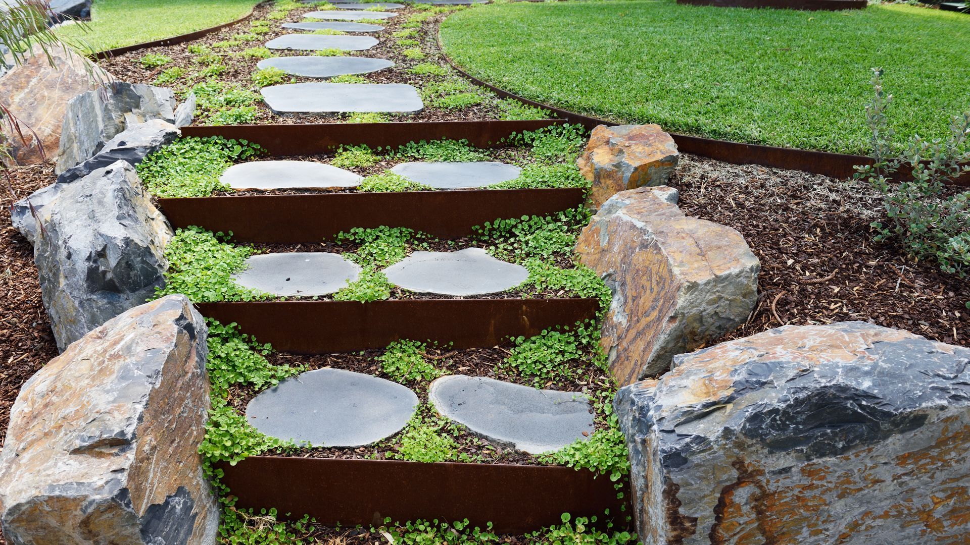 Stone steps in a garden, edged with rusty metal and large rocks, surrounded by greenery.