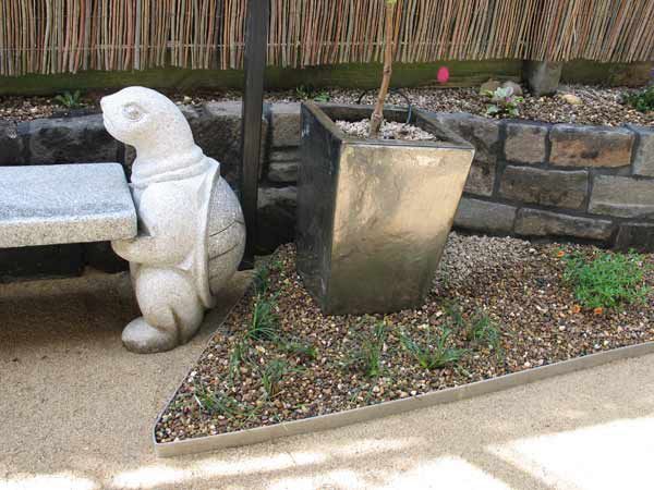 Stone squirrel statue holding a table, near a metallic planter and rock garden.