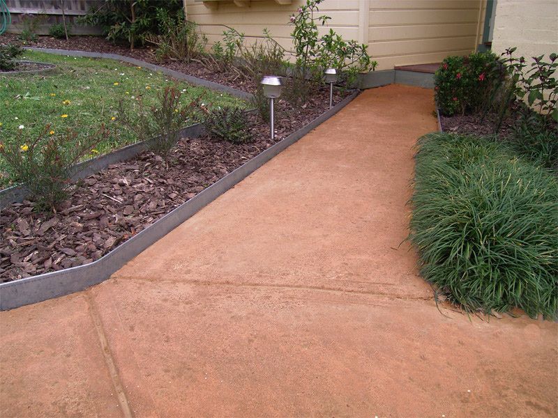 Red concrete pathway bordered by mulch and greenery, leading to a door with solar lights.