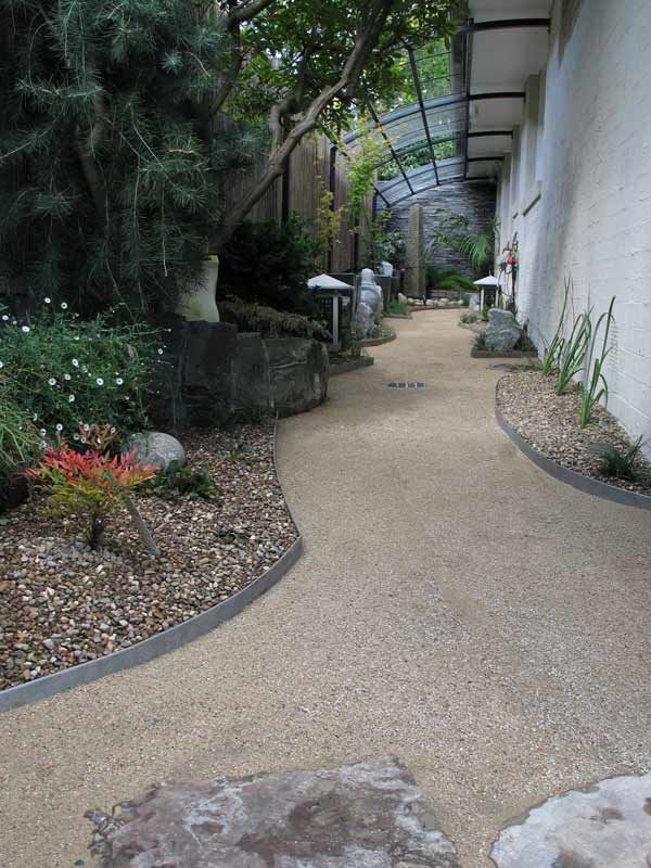 A winding gravel path in a garden with plants, stone lanterns, and a covered walkway.