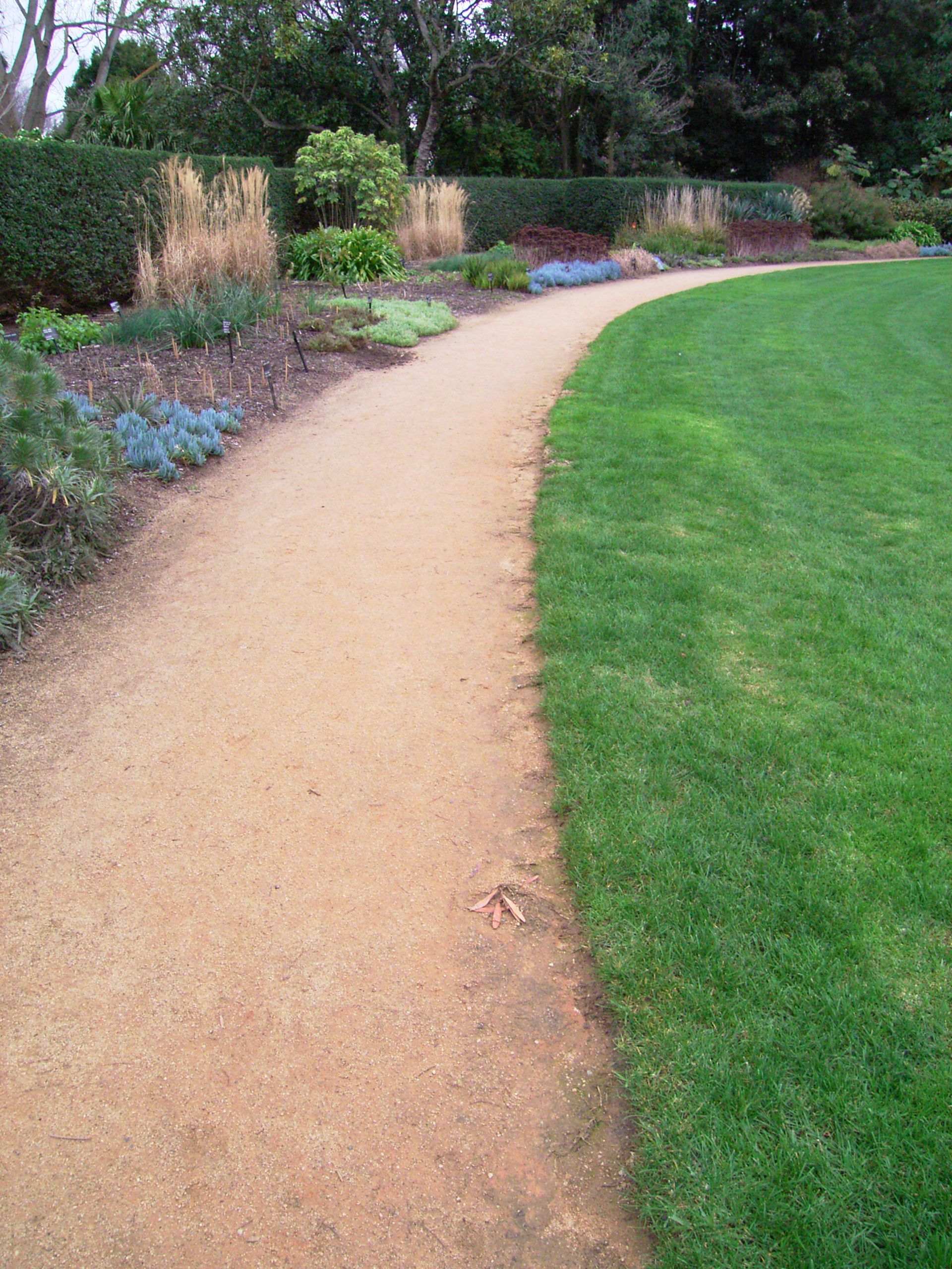 Curving tan path through a garden, bordered by green lawn and flowerbeds under a hedge.