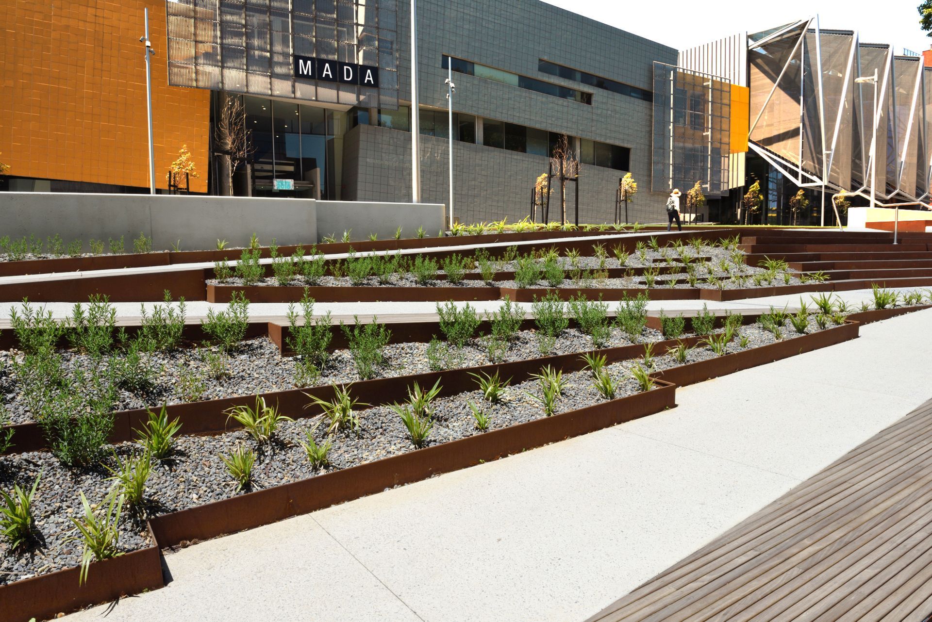 View of a modern building with tiered planters and a walkway. Building has orange and gray features.