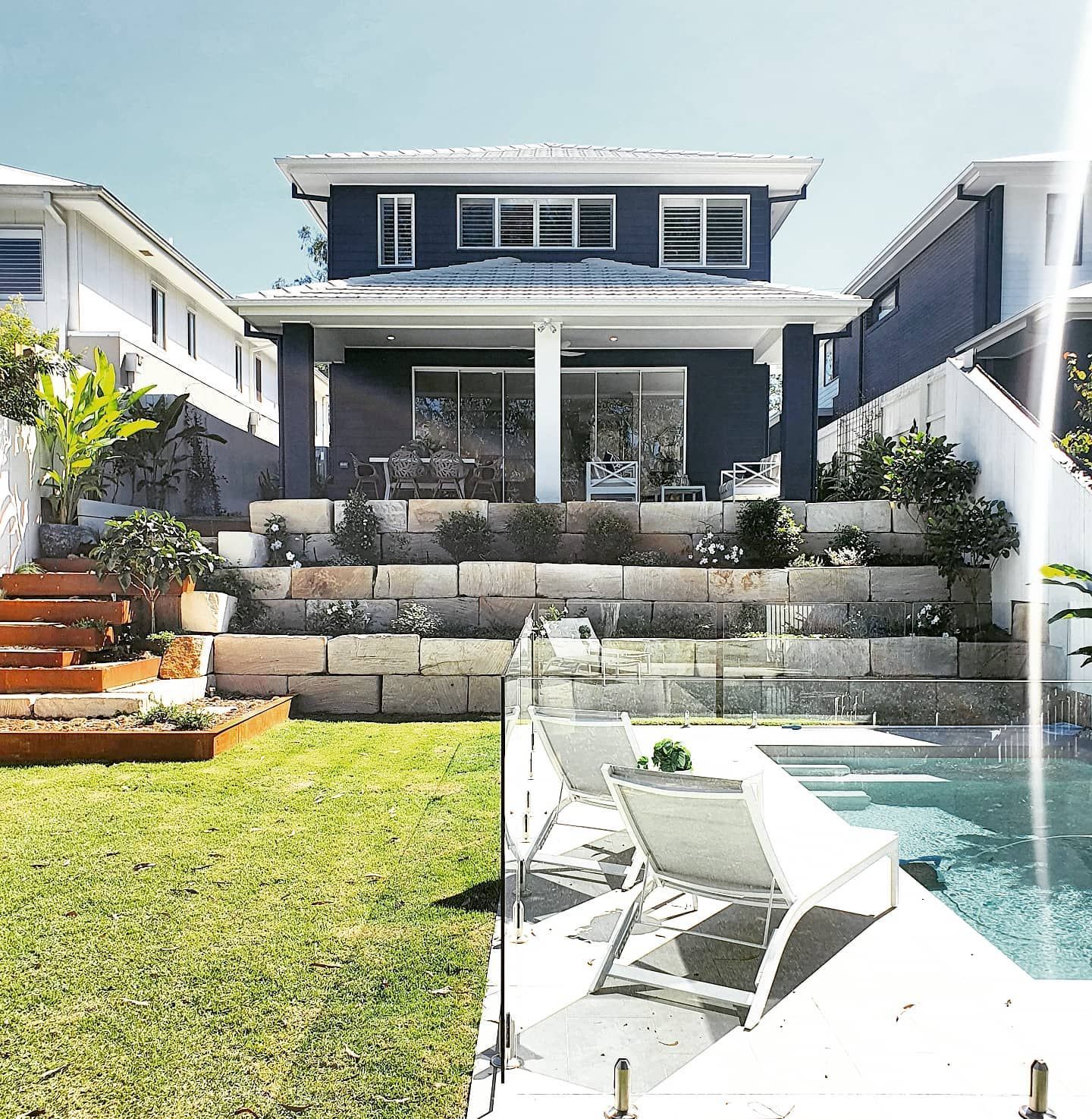 Two-story house with blue exterior, tiered landscaping, and a swimming pool in a sunny backyard.