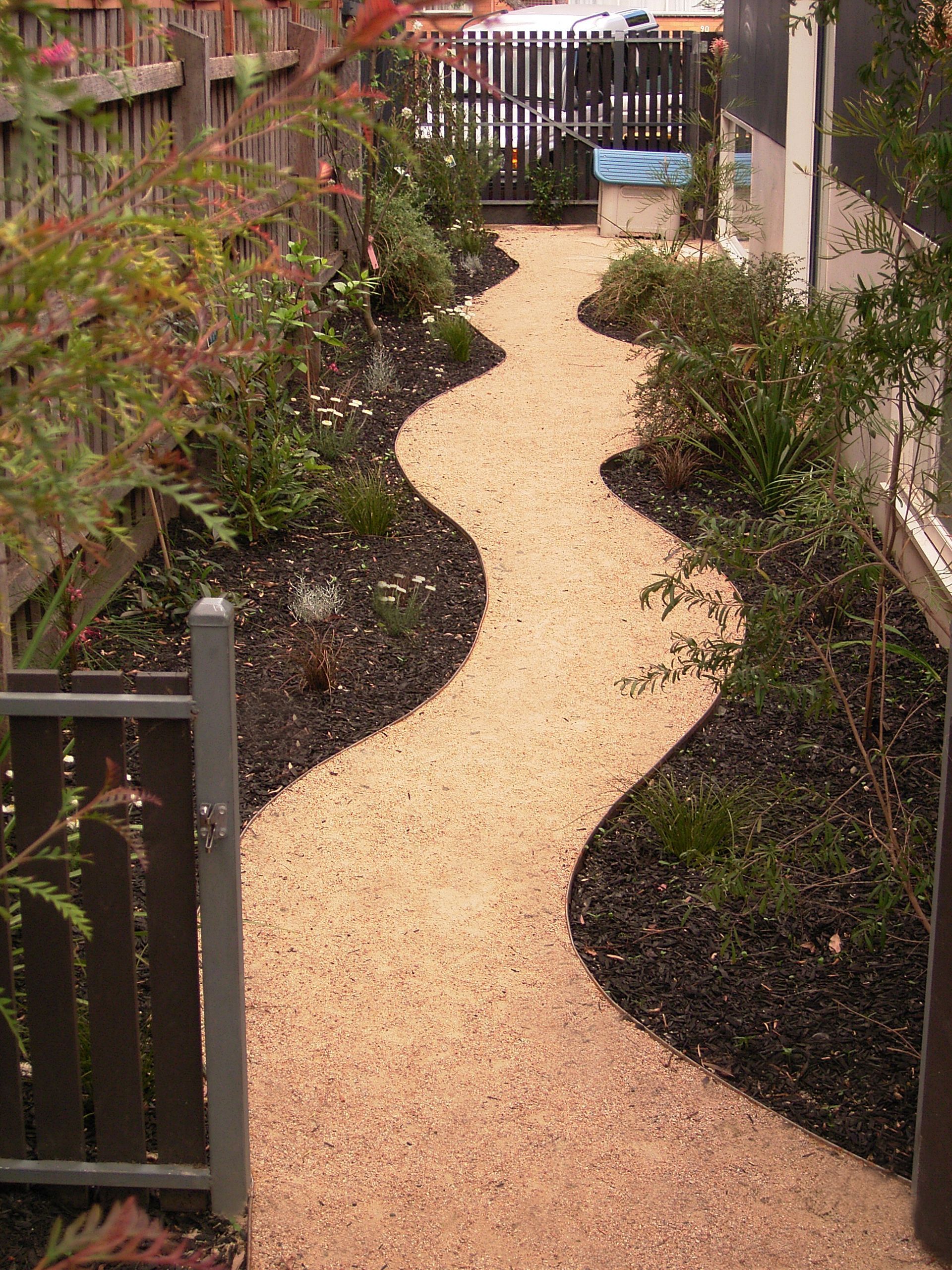 Curving gravel path through a garden with dark mulch and bordering plants, leading to a gate.