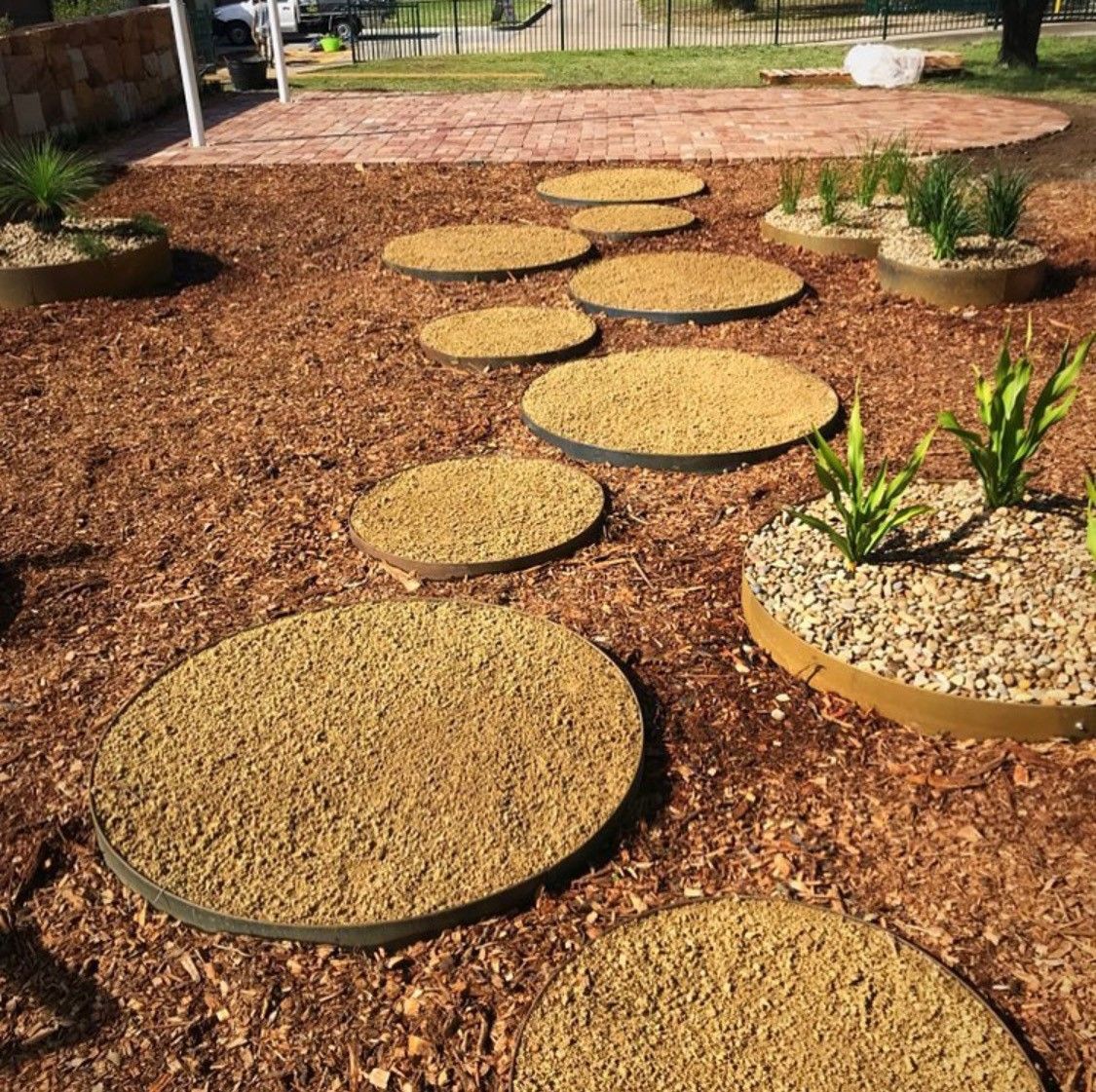 Circular stepping stones in a garden bed with plants, mulch, and a red brick patio in the background.