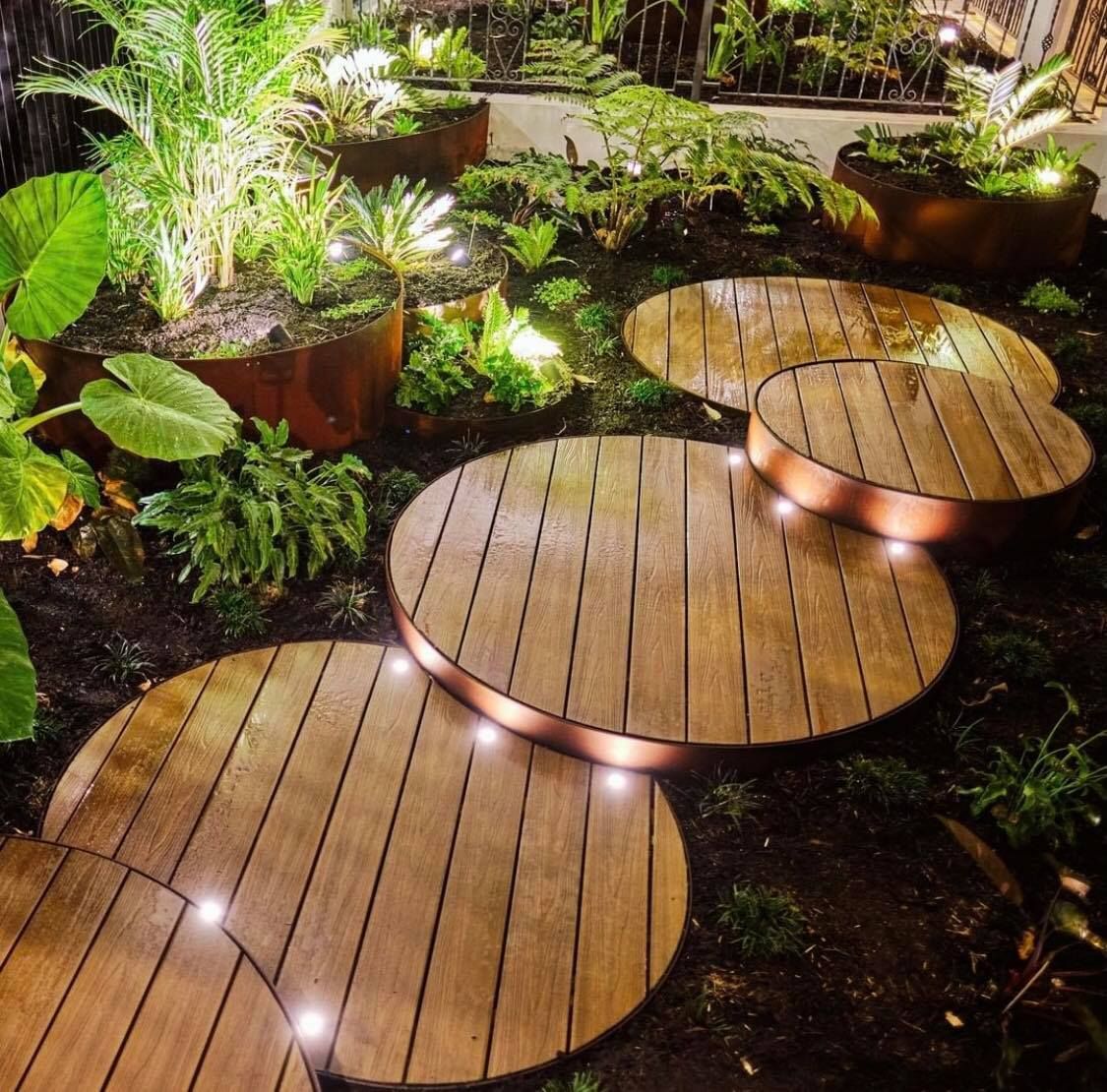 Wooden stepping stones illuminated in a garden with lush greenery and planters.