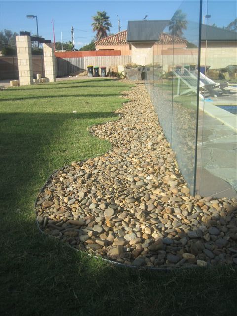 Lawn with a stone border next to a glass pool fence. Buildings and palm trees in the background. Sunny day.