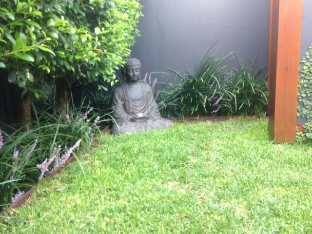 Buddha statue sits in a grassy garden, surrounded by plants and a dark gray wall.