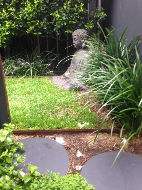 Buddha statue in a garden, seated on grass, surrounded by greenery and stone stepping-stones.