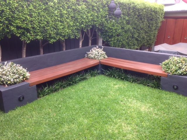 Corner wooden bench surrounded by dark gray walls and green grass, with a hedge background.