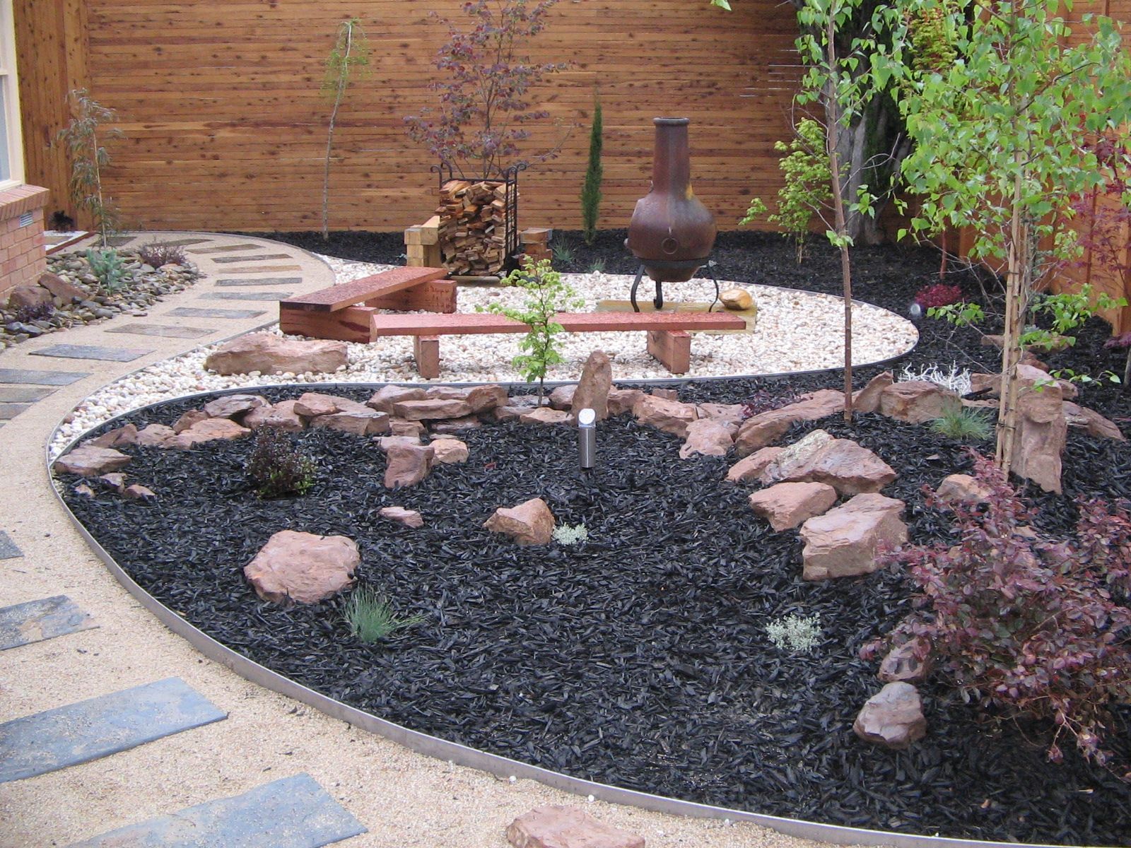 Zen garden with a curving path, rock features, and wood fence. Black mulch and white gravel are used.