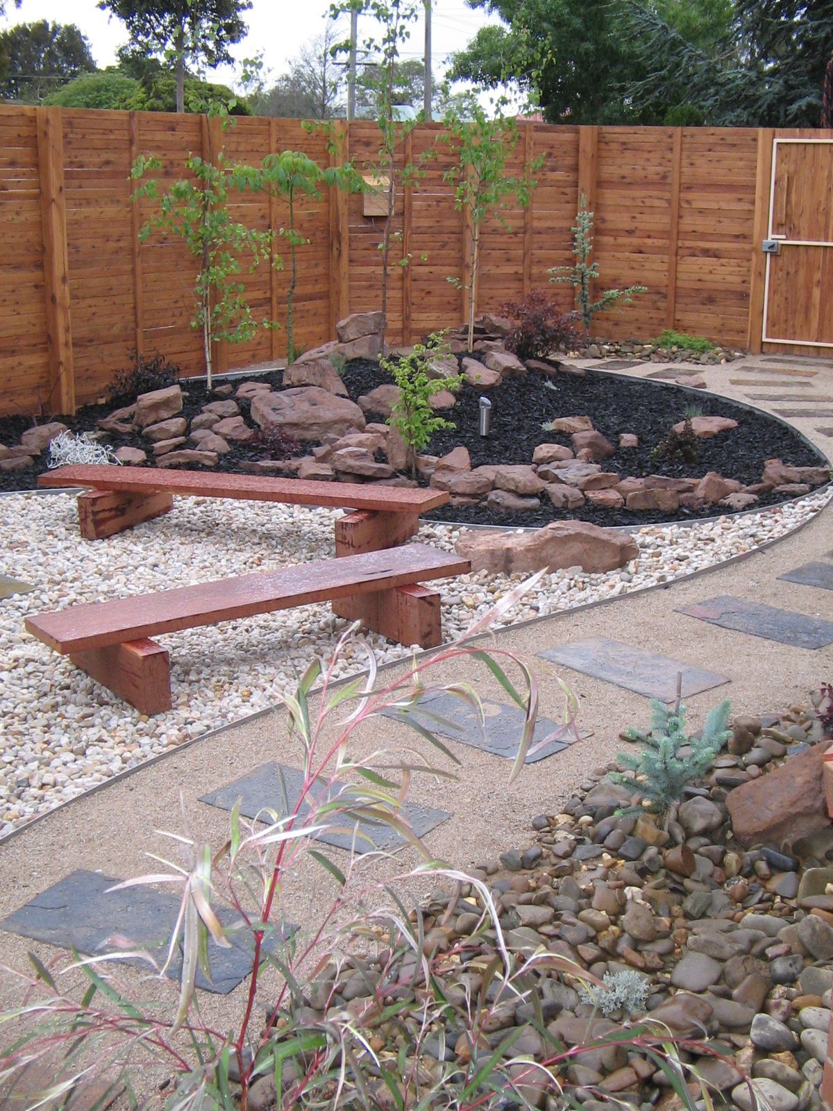 Wooden benches in a garden with rocks, trees, and a wooden fence.