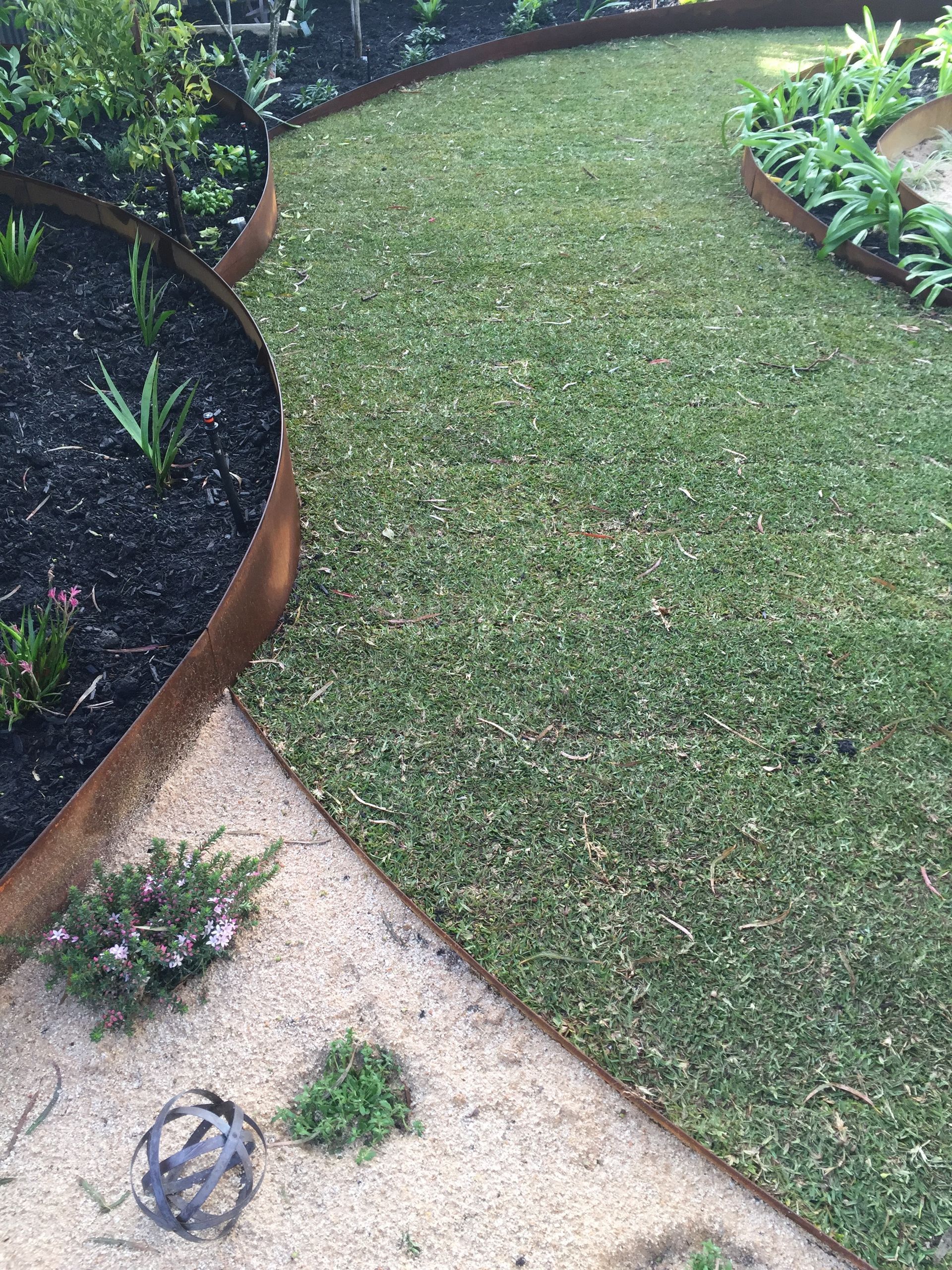 Curved path through garden with rusty metal edging. Lawn, mulch beds, gravel paths, and plants.