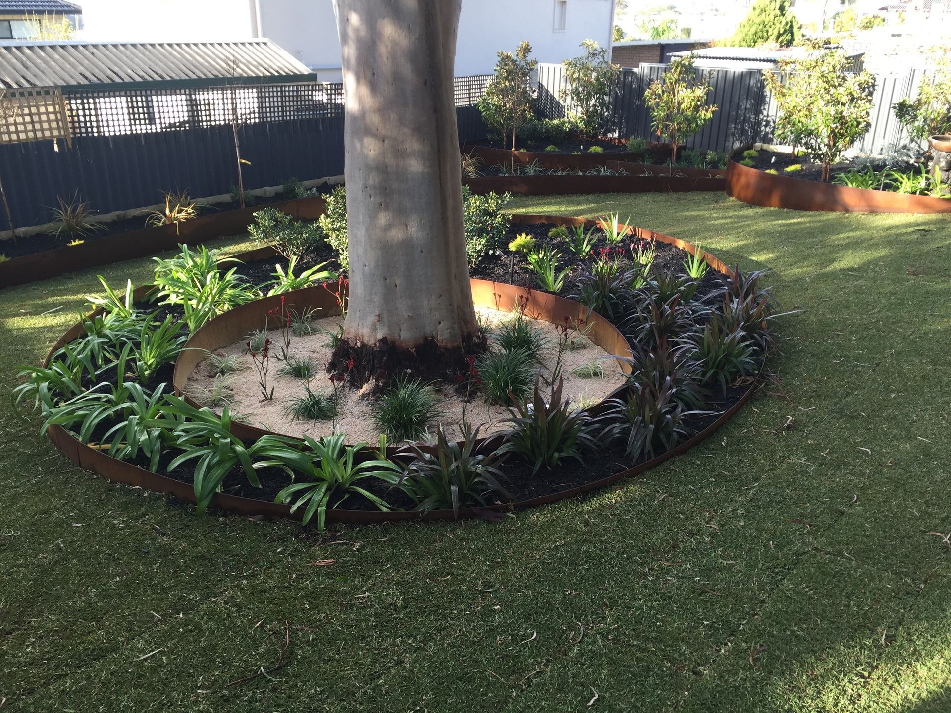 Circular garden bed around tree trunk with brown metal edging, green plants, and artificial turf.